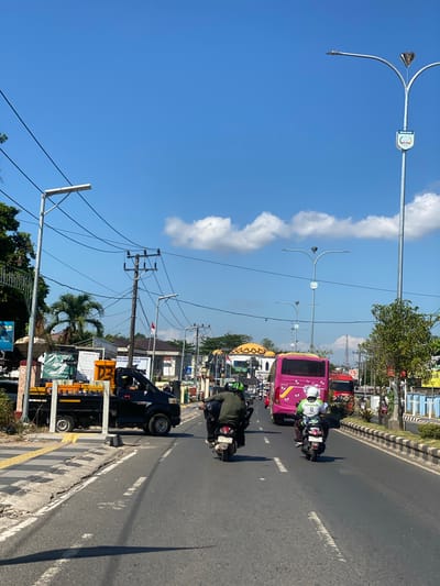 The image depicts a street scene in Jawa, Indonesia, under a clear blue sky with scattered clouds. The perspective is from a vehicle traveling on a paved road with visible lane markings. In the foreground, two motorcyclists are riding in the same direction. The first motorcyclist, wearing a green jacket and helmet, is ahead and slightly to the left. The second motorcyclist, wearing a white shirt with "Kawasaki" visible and a white helmet, is to the right and slightly behind.
In the middle ground, a black pickup truck with an orange payload is parked on the left side of the road, partially blocking a pedestrian crossing marked with yellow tactile paving. Further down the road, a bright pink bus is traveling away from the viewer, followed by a red bus. Several streetlights with distinctive decorative diamond-shaped elements are visible along the roadside, along with utility poles and power lines. Buildings line the right side of the street, some with distinct architectural features like a domed roof. Traffic signs are present, including a blue and white sign with the word "BANJAR" and an emblem. Vegetation, including palm trees and bushes, can be seen along the sides of the road and in front of some buildings. The overall impression is a busy urban environment on a sunny day.