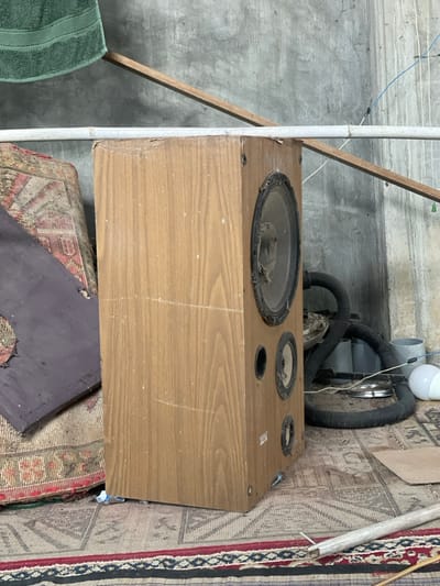 The image depicts an indoor scene in the city of Kumbang, Indonesia, focusing on a worn, light brown wooden speaker cabinet standing upright on a patterned rug.

The speaker cabinet features a prominent, dust-covered main driver with a torn or deteriorated cone and surround, positioned at the top. Below it are two smaller drivers, also dust-laden, with an open circular port (likely a bass reflex port) situated between them. The wood-grain finish of the cabinet exhibits numerous scratches, scuffs, and dust accumulation, with a chipped edge along the top. A horizontal white pipe, possibly PVC, rests on the top edge of the speaker.

To the left of the speaker, a stack of heavily soiled, patterned textiles or cushions is visible, partially obscured by a dark, flat, irregularly shaped object. The floor is covered by at least two layers of worn, patterned rugs or mats, displaying geometric and floral designs.

The background consists of a rough, grey concrete wall. To the right of the speaker, various objects are stored against the wall, including a dark, flexible corrugated hose (possibly from a vacuum cleaner), a metallic circular object (resembling a pot lid), a white incandescent light bulb, and light-colored plastic containers. Electrical wiring, including blue and white strands, is visible against the concrete wall. A diagonal wooden plank extends across the upper background, behind the white pipe. In the upper left background, a green towel hangs draped over an unseen support. A light-colored pole or stick lies on the rug in the lower right foreground. No people are present in the scene.