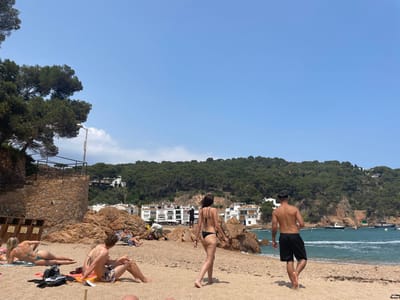 The image shows a beach scene in Tamariu, Spain (41.917, 3.207). There are three people on the beach, a man and two women. The man is wearing black swim trunks and is walking away from the camera. The woman on the left is laying on a towel, while the woman on the right is wearing a black bikini and walking towards the camera. There are several other people in the background, sitting on the beach and on a rock outcropping. The beach is sandy and there are rocky outcrops along the shoreline. The water is clear and blue, and there are boats visible in the distance. There is a large hill covered in trees in the background, with a small town visible behind the hill. The sky is blue and clear. The image appears to have been taken on a sunny summer day.