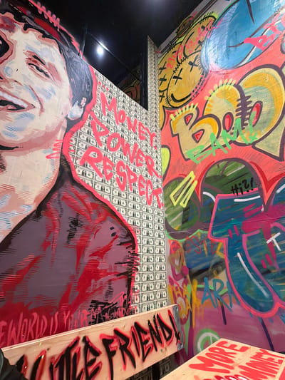 The image displays an interior space, likely a commercial establishment located in Bielefeld, Germany, characterized by extensive street art and graffiti. The left wall features a large, stylized pop-art mural of a smiling individual with closed eyes, rendered in light flesh tones, black, grey, and prominent fluorescent pink outlines. Below the portrait, partially visible text reads "THE WORLD IS YOURS" in red graffiti. Adjacent to the portrait, a panel or wall section is covered with a repeating pattern of US dollar bills, overlaid with prominent fluorescent pink graffiti spelling "MONEY POWER RESPECT" stacked vertically. The right wall is entirely covered in multi-layered, vibrant graffiti. Visible elements include a large yellow smiley face with "X" eyes and a stitched mouth, bubble letters in yellow and green possibly forming "BAM!", and various abstract shapes, tags, and letters in blue, pink, green, orange, and black, including the word "HI!" and "AR". In the foreground, two light-colored wooden structures are present: a bench-like object on the left with "LITTLE FRIENDS!" spray-painted in black with a red glow effect, and a partial wooden surface on the right, possibly a table, with "MORE MONEY" spray-painted in red. Overhead lighting is reflected on the right wall. The overall environment conveys an urban, artistic, and energetic atmosphere.