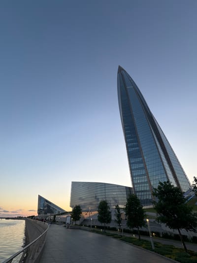 This is a wide-angle, low-angle shot of a waterfront promenade and modern buildings at dusk in the city of Alyumino, Russia. The sky is a clear gradient of pale blue to light yellow near the horizon. On the left, a calm body of water reflects the sky and the lights of the promenade. A curved concrete promenade lined with a metal railing runs along the waterfront. Several silhouetted figures are walking on the promenade.

Dominating the right side of the frame is a tall, modern skyscraper with a distinctive pointed, curved design, its glass facade reflecting the sky. To the left of this skyscraper are two other angular, glass-fronted buildings. The buildings are illuminated by artificial lights. Trees and landscaping with modern light fixtures line the promenade in front of the buildings. The scene captures a contemporary urban landscape by the water as day transitions into night.