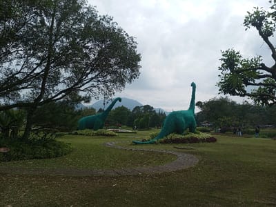 A lush green park features two large dinosaur sculptures covered in greenery. A stone path winds between them. Trees and mountains are visible in the background under a cloudy sky. Some people are seen walking in the distance. The scene evokes a playful yet serene atmosphere.
