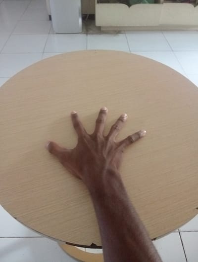 A left hand with fingers spread open rests on a light brown, circular table. The table has a wood-grain texture and is positioned on a white tiled floor. In the background, out of focus, are white cabinets and a white rectangular object that could be a refrigerator or trash can. The scene appears to be indoors, possibly in a kitchen or dining area. The lighting is bright, suggesting it is daytime. The location of Surabaya, East Java, is not discernible from the image itself.