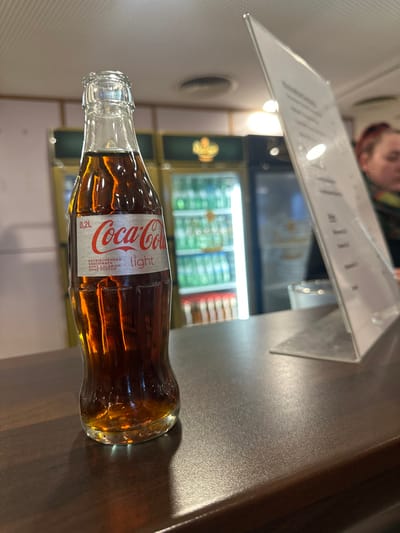 A Coca-Cola Light bottle, 0.2L, sits on a wooden table. The bottle is partially obscured by a reflection of the table. The bottle label is visible, showing the Coca-Cola Light branding and the words "Erfrischender Geschmack, mit Koffein, ohne Zucker." The table is partially visible with some of the surface in focus, and is situated in front of a blurry background. Behind the bottle are two refrigerators, partially visible, stocked with various beverages. The bottle is located in a public area. The coordinates provided indicate that this is in Bingen am Rhein, Germany.