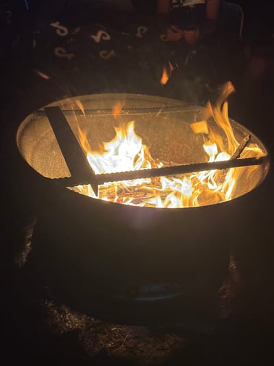 The image shows a close-up, eye-level view of a metal fire pit filled with burning logs and flames. The fire pit is a dark, circular metal structure with a rustic texture. Inside, brightly burning wood creates a vibrant display of orange, yellow, and white flames, with some smoke rising. Two metal rods are placed across the fire pit, supporting the burning logs. In the background, a dark blanket with a repeating pattern of white symbols is visible, along with the blurred figures of people sitting around the fire. The ground around the fire pit appears to be covered in dry leaves and debris. The overall scene depicts an outdoor campfire at night, likely in a rural or recreational setting, potentially in the city of Вязынскі сельскі Савет, Belarus, based on the prompt.