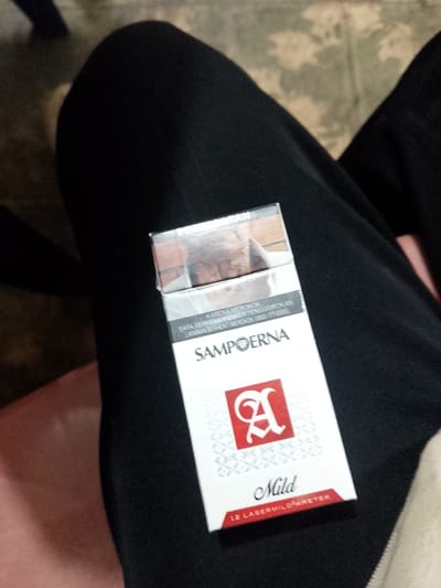 The image shows a close-up view of a pack of Sampoerna A Mild cigarettes held by a person whose legs are covered in black fabric. The cigarettes are resting on the person's lap, which is a dark, possibly navy or black, material. The background is out of focus, but appears to be a dark, indoor environment with some lighter areas suggesting furniture or clothing. There is no discernible information about the specific location in Lhokseumawe, Indonesia. The focus is on the cigarette pack, which features the brand name "SAMPPOERNA," the logo "A" in a red square, and the word "Mild" in script. The text "12 LASERMILD KRETEK" is also visible at the bottom of the pack. The image is taken from a slightly overhead angle.