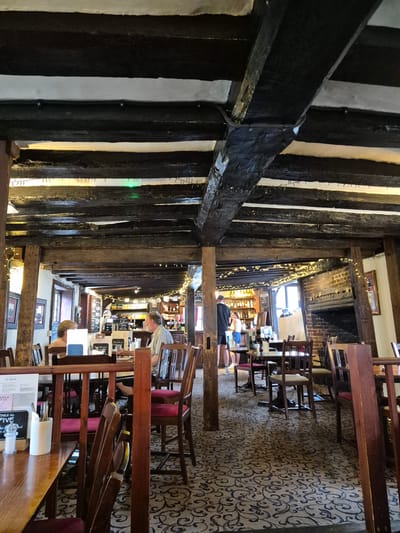 The image shows the interior of an old, possibly historic, pub or restaurant. The location is suggested to be in Babergh, United Kingdom. The building features exposed timber beams in a black-and-white Tudor style. Small fairy lights are strung along the beams.
People are present in the background, either standing at the bar or seated. A man with short grey hair and glasses is seated at a table. Another person is partly visible in the foreground, seated at a table. There are tables and chairs throughout the room. The floor has a patterned carpet. Menus, salt and pepper shakers, and other dining accessories are on the tables. The establishment appears to be open for business.