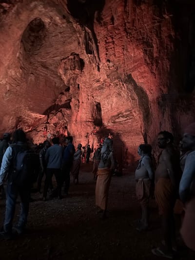 The image depicts a gathering of people inside a cavern, likely a tourist attraction or religious site, illuminated by red lighting. The cavern walls are rough and textured, with visible geological formations. A group of men, some appearing to be sadhus or ascetics with long beards and ash-smeared bodies, are present. They are wearing orange or white dhotis. Other individuals, possibly tourists, are also visible, some wearing modern clothing and backpacks. The ground is uneven and appears to be covered in gravel or dirt. The overall scene suggests a spiritual or cultural event taking place in a natural, cave-like environment. The location is identified as the Yaganti Caves in Bethamcherla, India.