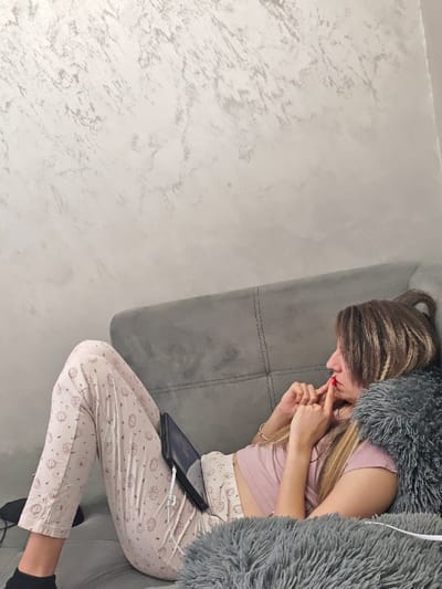 The scene is an indoor setting in Opaka, Bulgaria. An adult female is reclining on her left side on a gray fabric sofa. She has light brown hair with blonde highlights, partially tied up, and is wearing a short-sleeved light pink top and light-colored pajamas with a repeating pattern. Dark socks are visible on her left foot. Her left hand, featuring red nail polish, is positioned with fingers touching her lips. A dark-cased tablet, connected to white wired earphones, rests on her lap/midsection, displaying a screen. A dark gray, long-pile fluffy blanket or throw is draped over the sofa and partially covers her. The wall behind the sofa features a light, textured finish, resembling marble or stucco.