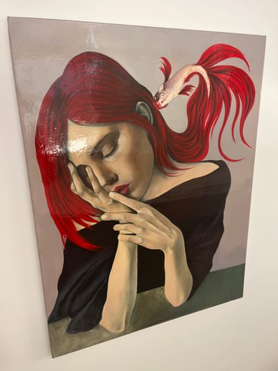 This image is a photograph of a vertically oriented painting, physically located in Galatina, Italy, displayed against a plain, light-colored wall. The painting depicts a woman from the chest up, with her eyes closed and her head slightly tilted downwards. She has vibrant, flowing red hair that extends upwards and to the right. Her left hand, with fingers slightly splayed, covers the lower part of her face, from just below the nose to her chin, and rests upon her right hand. Her right hand is positioned beneath her left, with fingers interlocked or crossed. She exhibits fair skin and visible red lips beneath her left hand. She wears a dark, off-the-shoulder garment. Above and to the right of the woman's head, a white-bodied fish with large, flowing red fins, resembling a Siamese fighting fish, is depicted. Its fins appear to be integrated with or emerging from the woman's red hair. The background of the painting is a solid light gray-lavender color in the upper half, transitioning to a dark green or brown horizontal band in the lower portion, suggesting a surface. The painting has a glossy, reflective finish, indicated by visible light reflections on its surface.