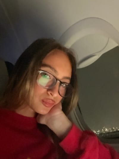 The image shows a young female inside an aircraft at night. The subject, positioned centrally, has long brown hair with lighter highlights and wears clear-framed glasses. She is dressed in a red long-sleeved garment, possibly a sweater or sweatshirt. Her chin rests on her left hand, and her lips are slightly puckered in a "duck face" expression as she looks directly towards the camera. A light source reflects prominently in her glasses. To the right, an aircraft window reveals blurred, distant city lights against a dark night sky, indicating the plane is airborne or on the ground at an airport. Above and behind her on the right, a curved white or light-grey panel, characteristic of an aircraft interior ceiling or wall, is visible. The immediate ground location, such as Sdot Dan Regional Council, Israel, cannot be determined from the visual information provided in the image.