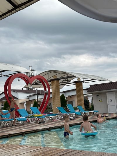 The image depicts an outdoor swimming pool area, likely part of a resort or leisure complex, in Yaroslavskaya, Russia. The scene is characterized by a cloudy sky and a shallow pool with clear water. In the foreground, a wooden deck surrounds the pool. Several children are visible in the water. One child in the foreground is wearing blue swim trunks and standing near the edge of the pool. Another child is in the middle of the pool, using a swim ring with a colorful pattern. Further back, another child is partially submerged in the water, holding a yellow water toy. To the right, a child is visible, also in the pool.
Along the edge of the pool, there are numerous bright blue lounge chairs. Some chairs have towels or personal items placed on them, suggesting recent use. A large, bright red heart sculpture, adorned with several disco balls, is a prominent feature in the background, positioned near the pool. Behind the heart sculpture, a series of buildings with white walls and dark roofs are visible, interspersed with greenery and trees. Covered seating areas with white canopies are present over sections of the lounge chairs and walkways. A small building, possibly a changing room or facility, with a white door and a sign on the wall, is located to the right of the image. The overall atmosphere suggests a recreational setting on an overcast day.