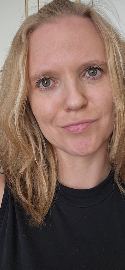 This is a close-up, chest-up portrait of a fair-skinned Caucasian woman with blonde hair, taken indoors in a neutral-toned environment, possibly the city of Ovoshchnoy, Russia. She is looking slightly off-camera with a faint smile. Her hair is parted to her left and falls in waves around her face and shoulders. She is wearing a black sleeveless top. The background is out of focus, but appears to have a vertical element, possibly a light-colored decorative screen or part of a furniture item. The lighting is soft and natural, illuminating her face evenly.