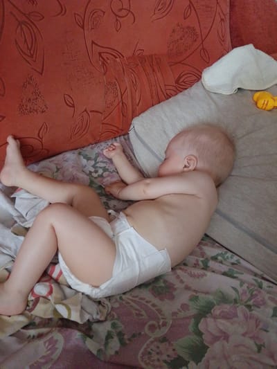 In this close-up shot, a baby is sleeping on its side. The baby is wearing a white diaper and has fair skin and light blonde hair. Its head is resting on a light gray pillow, and its body is positioned on a patterned bedspread with floral designs in shades of pink, green, and purple. A red, textured cushion is visible in the background, suggesting the baby is on a couch or bed. A white cloth is tucked behind the baby's head, and a small yellow toy, possibly a duck, is partially visible on the pillow to the right. The lighting suggests it might be daytime, and the overall scene conveys a sense of peace and rest. No text is visible in the image. The location context is Birsk, Russia.