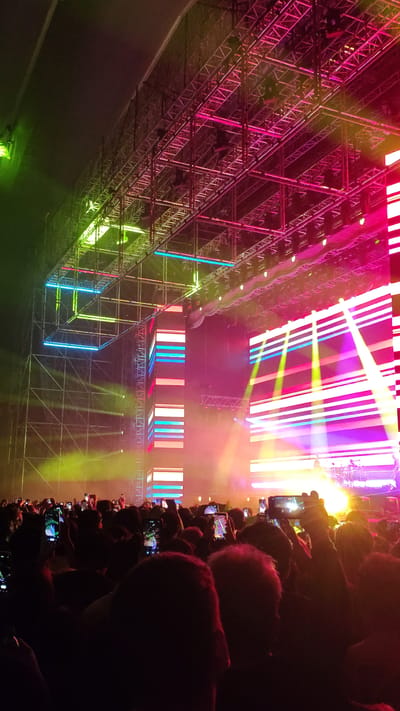 The image depicts a concert or performance scene, likely indoors, taken from the perspective of someone in the audience. The foreground is dominated by the silhouetted heads and hands of audience members, many holding up smartphones to record the event. Beyond the audience, a complex metal structure serves as a framework for lighting and visual displays. The structure is illuminated in vibrant colors, including pink, purple, green, blue, and yellow. Various light beams and panels add to the visual spectacle. Based on the lighting and set-up, the scene appears to be an electronic music event or a similar type of performance. There is no definitive information in the image to identify its location as San Miguel, Peru.