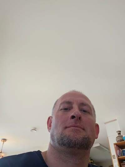 A close-up, low-angle selfie of a bald, middle-aged man with a short, graying beard is taken from a slightly upward perspective. He is wearing a dark blue t-shirt. The background consists of a white ceiling with a smoke detector and a light fixture visible. To the right, a portion of a bookshelf with decorative items, including a striped vase, is in view. The setting appears to be an indoor residential environment. There is no information available about the location in Smålandsstenar, Sweden.