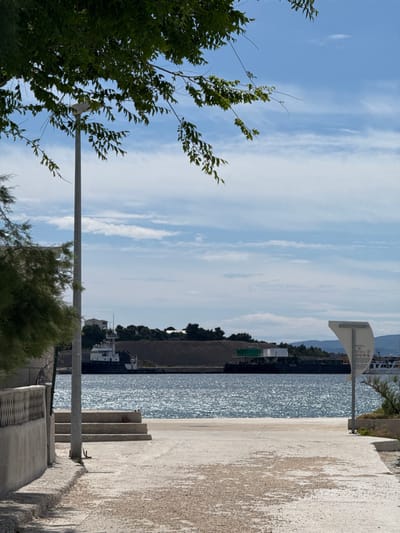The image shows a view from a paved pathway towards the sea in Dugi Rat, Croatia (latitude: 43.446, longitude: 16.634). A tall, thin light pole is on the left side of the path. There are white cement steps on the left side of the path leading up to a small concrete wall. A tree with thick, dark green leaves frames the scene on the left side of the image, partially obscuring a building with a white fence in the background. The path leads to the edge of the sea, where the water is clear and blue with gentle waves. In the distance, there is a small, white boat docked at the shore, and a larger dark-colored barge is moored. A green and white structure is on the barge. A white sign with black lettering is on the right side of the path. The sky is clear and blue with white clouds. The photo was taken on a sunny day.