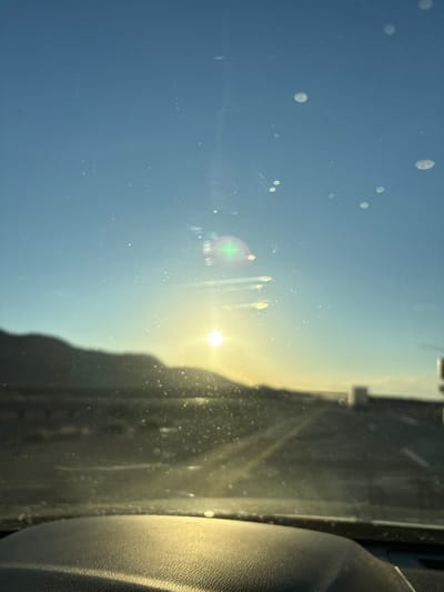 This image captures a view through a dusty windshield during what appears to be a bright, sunny day, likely late afternoon given the low angle of the sun. The sun itself is a prominent element, casting a warm, bright glow and creating lens flare effects that streak across the glass. The sky is a clear, pale blue, transitioning to a lighter, almost white hue near the horizon.

The foreground is dominated by the windshield itself, which is visibly dirty, with numerous white, circular smudges and smaller specks scattered across its surface. These imperfections obscure the view to some extent, creating a hazy, dreamlike quality. Through the windshield, the suggestion of a landscape is visible, including a distant, silhouetted landmass that could be hills or mountains, and some linear elements that might represent roads or structures. The right side of the frame shows a dark, textured surface, possibly the interior of a vehicle, such as a car seat or dashboard, with a coarse, leathery appearance. This detail grounds the scene, indicating the perspective is from within a vehicle.

The overall impression is one of travel or a road trip. The bright sunlight and clear sky suggest fair weather, and the presence of dust on the windshield points to a dry, possibly arid location. The time of day is further emphasized by the long shadows and the warm, golden light bathing the scene. There are no people or explicit text visible in the image. The mood is serene and contemplative, evoking the feeling of looking out at the world from a personal space while on the move. The location context of Yermo, USA, which is in a desert region, aligns with the dry, dusty appearance of the windshield.