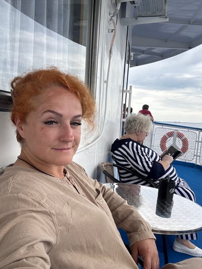 A woman with vibrant red hair and fair skin is captured in a selfie, looking directly at the camera with a slight smile and her head tilted. Her eyes are adorned with false eyelashes, and there are visible marks on her cheek and forehead. She is wearing a light brown button-up shirt. To her right, further back, an older woman with curly white hair is seated, wearing a navy and white striped top and dark pants, engrossed in her phone. She is seated at a glass-topped table with a black mug on it. The setting appears to be an outdoor deck of a vessel, likely a ferry, with a blue deck, white railings, and a life preserver visible in the background. The sky is overcast. The location is identified as Krasny Bor, Russia.