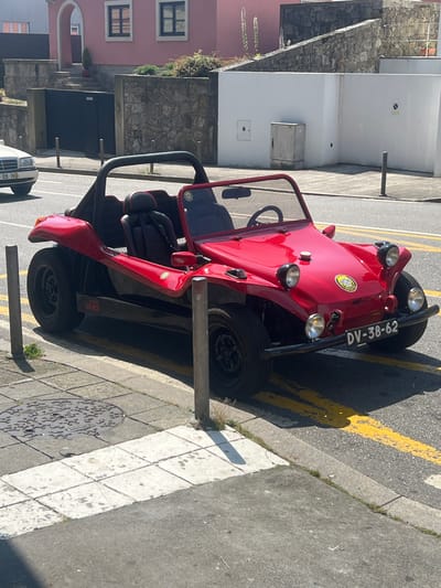 In the city of Viana do Castelo, Portugal, a bright red dune buggy is parked on a street. The buggy has a black soft top and black seats. On the side of the buggy, the words "TEAM PARIS" are visible in black lettering. The license plate reads "DV-38-62". The buggy is parked on the right side of the street, next to a sidewalk with a manhole cover. In the background, there is a pink building with arched windows, a stone wall, and a white building. Yellow lines are painted on the asphalt road.