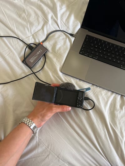 The image shows a close-up, overhead view of a person's arm and hand holding a black Sony camera with its screen flipped open. The camera is connected by a black cable to an external drive, which is also connected to a silver laptop. The laptop is open and positioned on a wrinkled white bedspread. The arm is wearing a silver watch. The scene is set on a bed, likely in a bedroom. The location is not Borowa, Poland.