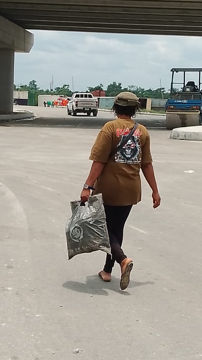 An outdoor shot depicts a person with their back turned to the camera, walking on a concrete ground beneath an overpass. The person wears a brown t-shirt with a skull graphic on the back, dark pants, and flip-flops. They are carrying a gray plastic bag in their left hand and have a hat on their head. A shoulder strap is visible across their back.
In the background, a white Toyota pickup truck is driving away, with a group of people wearing orange vests standing near it. To the right of the truck is a construction vehicle, a blue vibratory roller. The area seems to be an urban construction site.
Given the Effurun location, this suggests the image was taken somewhere in Effurun, Nigeria.