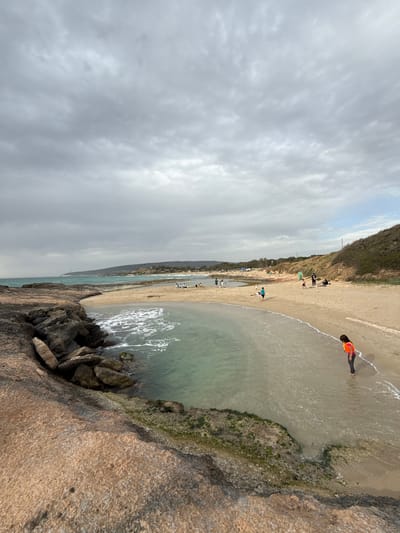 The photograph shows a beach on the coast of Nahariah, Israel. Several people are visible along the shoreline, including an individual in an orange life vest standing in the shallow water. The beach is sandy, with a ridge of vegetation and soil lining the edge of the coast. The foreground shows rocky terrain with seaweed and moss. The sky is overcast with heavy clouds. The water is calm and clear, revealing a turquoise hue.