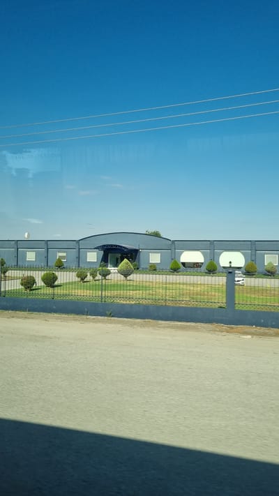 A long, low, grey building with a curved central section is visible under a clear blue sky. The building has multiple rectangular windows and two large circular windows on the right side. A dark blue awning extends over the entrance. In front of the building is a manicured lawn with several cone-shaped evergreen bushes. A dark grey metal fence encloses the property, and behind the fence is a grey asphalt surface. Power lines are visible in the upper portion of the image.