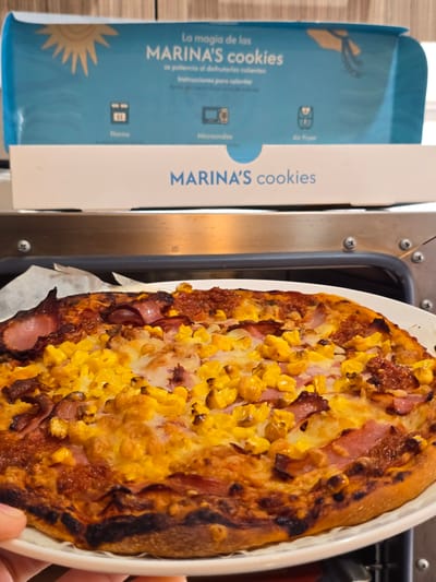 The image shows a pizza with corn and ham toppings next to a box of "Marina's Cookies". The box mentions heating instructions for oven, microwave, and air fryer. The photograph appears to be taken indoors, possibly in a kitchen. The image does not contain information about the city of La Florida, Chile.
