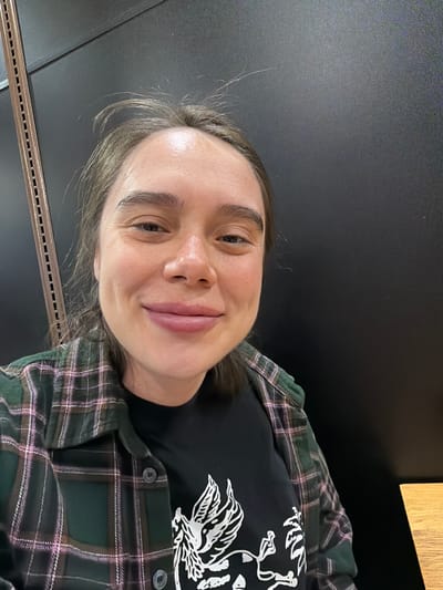 A person with light skin, brown hair, and brown eyes smiles slightly towards the camera. They are wearing a black t-shirt with a white design and a green and purple plaid flannel shirt. The background appears to be a dark gray or black surface, potentially a wall or table. A thin, metallic strip is visible along the bottom edge of the frame. The image was taken indoors. There is no verifiable information that this image was taken in Niles, United States of America.