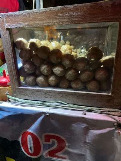 The image displays a close-up view of a street food vendor's setup, presumed to be in Kauman, Indonesia. The primary subject is a rectangular display case constructed from dark brown, weathered wood framing clear glass panels. Inside, hundreds of light brown, spherical food items, consistent with meatballs (bakso), are densely stacked and partially illuminated from within, creating reflections on the glass. Below the display case, a light-colored banner or tarp is affixed, showing a partial red numeral "02". To the upper left, the upper arm of an individual, wearing a black shirt with a red sleeve or accent, is partially visible. On the left side of the frame, adjacent to the display, a red plastic squeeze bottle and a yellow container are partially visible. The scene is dimly lit, suggesting an evening or nighttime setting.