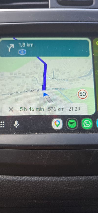 The image displays a navigation screen, likely from a car's infotainment system, showing a map with a route highlighted in blue. The screen indicates a distance of 1.8 km to the next turn, with the number 8 in a blue shield, suggesting a highway or main road. The route appears to be heading towards "Zum Kruckelsberg." Below the map, a journey summary shows an estimated time of 5 hours and 46 minutes and a total distance of 576 km, with an arrival time of 21:29. A speed limit sign for 50 km/h is visible on the right side of the map. At the bottom of the screen, icons for Google Maps, Spotify, and WhatsApp are displayed, along with a grid icon and a microphone icon, indicating potential app shortcuts. The screen is mounted in a car dashboard, with the texture of the dashboard visible. The context of Schwalbach, Germany is provided as the location.