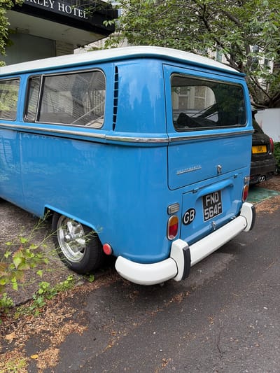 The image features a classic light blue and white Volkswagen Type 2 camper van, seen from its rear-right side. The van is parked on an asphalt surface that appears damp, possibly from recent rain. Its bodywork shows a two-tone scheme with a white roof and lower section, and a light blue main body. Chrome trim highlights the windows and runs along the side of the vehicle.

The rear of the van displays classic round taillights and a prominent white, curved bumper with black rubber accents. A black British license plate, clearly visible, reads "FND 564F," accompanied by a circular "GB" sticker. The word "Volkswagen" is embossed in chrome script on the engine cover below the rear window.

Behind the van, a dark-colored SUV is partially visible, showing a yellow UK license plate with the text "1AVL." To the left of the van, a building stands with a black sign that has white lettering. The visible portion of the sign reads "LEY HOTEL," suggesting a hotel establishment, likely "HARLEY HOTEL" or similar given the Harrogate context. Green foliage and tree branches partially obscure the building and van, with scattered fallen leaves on the ground. The scene is illuminated by natural daylight, consistent with an overcast or post-rain condition.