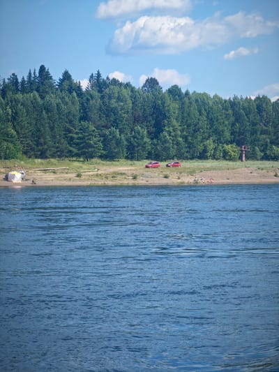 The image depicts a sunny day at a body of water, likely a river or lake, in the vicinity of Abalakovo, Russia. In the foreground, the water's surface is rippled, reflecting the blue sky and scattered white clouds.
Along the shore in the middle ground, a sandy bank is visible, backed by a dense forest of evergreen trees. On the sandy bank, several objects and individuals are present. Two red cars are parked on the grass verge near the trees. Closer to the water, a pop-up tent with white and orange panels is set up, with a single person sitting nearby. Further along the beach, a small group of people are gathered on blankets, some appearing to be sunbathing or relaxing. To the far right of the sandy area, a red metal tower structure stands. The overall environment suggests a recreational area by the water.