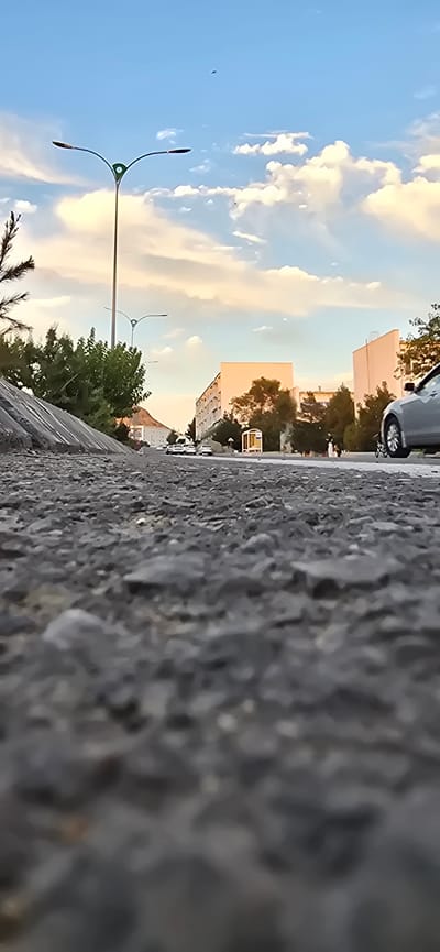 An eye-level, low-angle shot captures a street scene in Türkmenbaşy, Turkmenistan, emphasizing the textured asphalt in the foreground. To the left, a concrete barrier slopes upwards, topped by lush green trees and a pine tree. A modern streetlamp with two lights and a decorative green emblem stands tall against a bright blue sky dotted with white clouds, some tinged with the warm hues of sunset. In the middle ground, several cars are parked and in motion on the road, with buildings lining the right side of the street. The buildings are modern, multi-story structures, some appearing residential or commercial, with visible windows and balconies. A small, transparent glass kiosk or bus stop is situated near the buildings on the right. A light-colored car is prominently in motion on the right side of the frame, partially obscuring the view of other vehicles and the buildings behind it. The sky transitions to warmer tones towards the horizon, suggesting late afternoon or early evening.