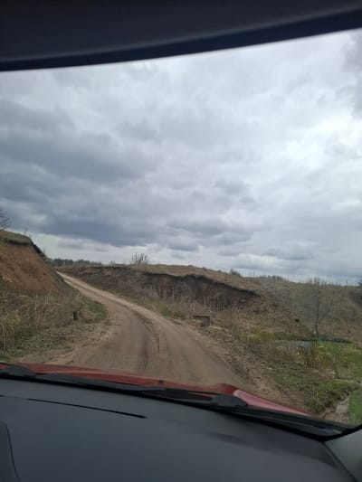 The image, taken from inside a vehicle looking out the front passenger side, captures a rural or natural landscape under an overcast sky. The view is tilted, suggesting the photo was taken from a vehicle on an incline or with an unlevel horizon.

The upper portion of the frame is dominated by a vast, grey, cloudy sky, indicating cloudy weather. Below the sky, a dirt road, brown and uneven, curves through a landscape of rolling hills and embankments. The terrain is covered in dry, sparse vegetation, consisting mainly of light brown grass, leafless bushes, and a few bare trees, suggesting late winter or early spring. The hillsides show variations in elevation and texture, with some areas appearing as steep cuts or natural slopes.

Parts of a red vehicle are visible on the right side of the image, including the front pillar and a section of the hood or fender, along with a dark grey interior element or window frame. A similar dark grey edge is present on the far left. The vehicle appears to be either traveling or parked on this unpaved road. The diffused light suggests it is daytime. There are no people visible, nor any discernible text in the image. The setting is identified as Yablonovo, Russia.