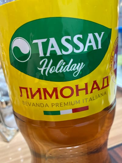 The image shows a close-up of a yellow plastic bottle with a green and white label. The label features the brand name "TASSAY" in large white letters, followed by "Holiday" in cursive. Below this, in red Cyrillic letters, is the word "ЛИМОНАД" (LIMONAD), which means lemonade. Underneath, in smaller white letters, it reads "BEVANDA PREMIUM ITALIANA", indicating it is a premium Italian beverage. The bottle appears to be partially filled with a brown liquid. There are also faint hints of other objects in the background, including what looks like a glass object to the left and some light-colored specks on a wooden surface below the bottle. The context of the location in the city of Әуезов ауылдық округі, Kazakhstan is not directly visible in the image.