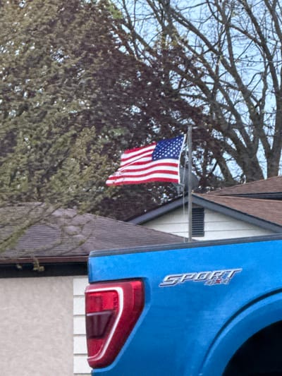 The image depicts an American flag hanging from a house in Grove City, Ohio, United States. The flag is partially obscured by tree branches. The tail light and part of the body of a blue vehicle, bearing the "Sport" emblem, are visible on the right side of the image. The house has brown trim and white siding.