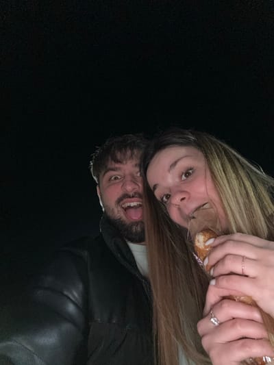 Two individuals, one male and one female, are depicted in a close-up, low-light photograph taken in Frătești, Romania. The background is uniformly dark, suggesting a nighttime or dim environment. The male, positioned on the left, has dark hair and a beard, wearing a dark puffer jacket over a light-colored garment. He exhibits an exaggerated facial expression with wide eyes and his tongue extended. The female, positioned on the right, has long light-brown hair and wears a ring on her left ring finger and a thin bracelet on her left wrist. She is holding a cylindrical food item, possibly a pastry, partially wrapped in clear plastic, to her mouth. A dark filling is visible at the point where she is engaging with the food. Both individuals are looking directly towards the camera.