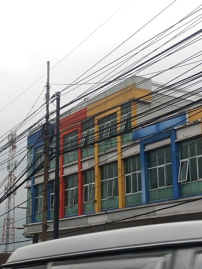 The scene depicts an urban street view in Mandandawela, Sri Lanka, under an overcast sky. A multi-story building dominates the midground, featuring a facade with alternating vertical sections of blue, red, and yellow. Numerous rectangular windows with white frames and green-tinted glass are visible across at least two floors, several of which are partially open, tilting outwards. A dense network of black electrical or communication wires spans horizontally and diagonally across the upper and middle portions of the image, partially obscuring the building and the sky. Two utility poles, one thick and dark, the other thinner and lighter, stand in the left-center, heavily laden with these wires. In the distant background on the far left, a tall, red and white lattice communication tower is visible. The top of a silver or grey vehicle occupies the bottom quarter of the frame, suggesting the photo was taken from or over a vehicle. No individuals or specific actions are depicted.