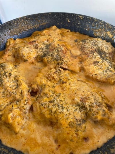 A close-up, overhead shot shows pieces of meat, possibly chicken, simmering in a creamy, orange sauce in a black frying pan. Dried herbs, likely dill, are sprinkled over the meat and sauce. The pan is dark and appears to be seasoned, with some food residue visible on its inner rim. The lighting is bright, creating a warm, inviting glow over the dish. The setting is likely a kitchen in Skala Avdiron, Greece, though no specific landmarks or environmental details confirm this location.