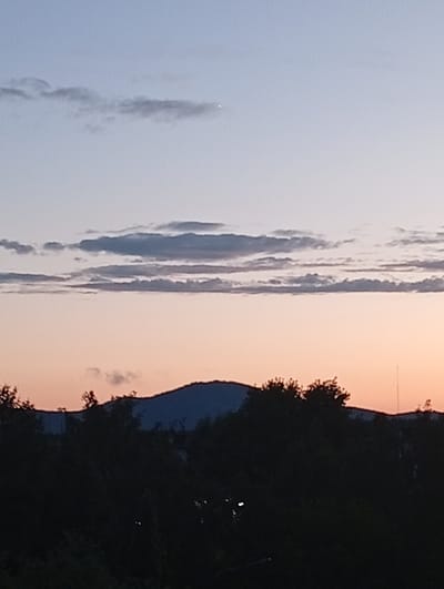 The image captures a twilight scene over Kyshtym, Russia, with a clear horizon dominated by a silhouette of trees and a mountain range. The sky transitions from a soft blue at the top to a warm, peachy-orange hue closer to the horizon, indicating either sunrise or sunset. Scattered across the upper portion of the sky are wispy, dark clouds, adding texture to the vast expanse. A single, bright star or planet is visible in the upper left quadrant of the sky. The foreground is filled with the dark silhouettes of dense foliage and trees, obscuring any finer details of the landscape below. The mountain range in the background is also a dark silhouette against the colorful sky, its peak gently undulating. The overall mood is serene and tranquil, typical of a quiet evening or early morning. There is no visible text in the image.