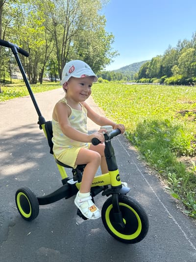 A young child wearing a white hat with strawberry patterns and a yellow tank top with shell designs is riding a yellow and black tricycle. The child is smiling and looking upwards, appearing happy. They are wearing white sandals with cartoon animal designs and yellow socks. The tricycle has a black handle and a push bar, suggesting it might also be parent-controlled. The child is on an asphalt path, likely in a park or recreational area. Green grass and trees line the path, and a hill or mountain is visible in the background, partially covered in trees. The sky is clear and blue. The overall scene suggests a sunny day. The word "MOOVEE." is visible on the tricycle's frame. The context provided indicates the location is Ustroń, Poland.