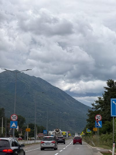 The image captures a tilted view of a busy two-lane road set against a dramatic mountainous backdrop under a cloudy sky, likely near Danilovgrad, Montenegro.

The road, appearing to run along the base of a mountain, is occupied by several vehicles. In the foreground and mid-ground, a dark grey SUV/hatchback, a silver Volkswagen SUV/hatchback (with the Montenegrin license plate "HN BT052"), and a red car are visible, indicating active traffic.

Numerous road signs line the route: multiple circular "50" km/h speed limit signs, a blue triangular sign with an upward-pointing arrow (indicating a hump or bridge), a "STOP" sign, and several pedestrian crossing signs. A yellow directional sign points to "Plana Bileca." A large blue directional sign with a white arrow pointing left is prominent at the top right. Streetlights or utility poles are spaced along the road.

To the left of the road, a large, tree-covered mountain dominates the landscape, its slopes densely forested with green foliage. The sky above is heavily overcast, filled with thick, grey and white clouds. The weather appears cloudy, and it is daytime. The scene conveys a sense of a routine commute or travel through a rugged, natural environment.