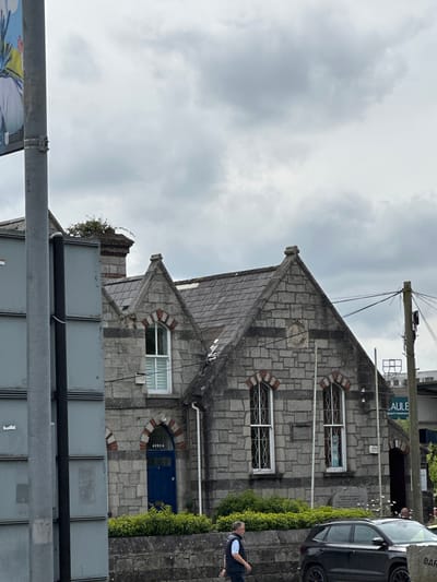 The image captures a cloudy day scene in Carlow, Ireland, focusing on the stone facade of a building with a distinctive pitched roof. The building features several arched windows with intricate metal grilles and white frames. A bright blue door is visible, with the word "AVOCA" written above it in white lettering. Lush green bushes line the front of the building. To the right, a utility pole with a network of wires stands prominently. In the foreground, a dark-colored car is parked on the street, and a man in a dark vest and white shirt is walking away from the camera towards the left. A large, grey, rectangular structure, possibly a billboard or signage, is partially visible on the left side of the image, obscuring part of the building. The overall impression is one of a historic building situated within an urban environment.