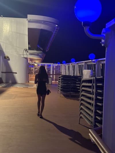 A full-body shot shows a woman standing on a deck at night. The image is oriented sideways, with the woman appearing to walk toward the right. She is wearing a dark, short-sleeved dress and her hair is dark and shoulder-length. She is also carrying a small black bag over her shoulder. The woman's shadow is cast long on the deck to her right.
The scene appears to be on the deck of a cruise ship or a similar vessel. In the background to the left, there is a white structure with an arched opening, possibly a part of the ship's superstructure. Further left, a large, bright blue light globe is visible, attached to a curved metal arm. Several other smaller blue lights are strung along the edge of the deck, creating a festive atmosphere.
To the right of the woman, rows of stacked chairs are visible, suggesting an outdoor seating area. The deck itself is a light brown color. The sky is dark, indicating it is nighttime. The lighting from the blue globes and other ambient lights casts a warm glow on the deck and creates distinct shadows. The overall mood seems relaxed and enjoyable, perhaps during a vacation or a special event. There is no visible text in the image.