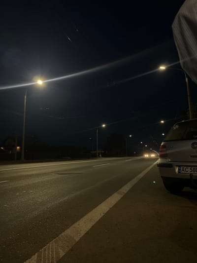 A wide, paved road stretches into the distance under a dark night sky. Streetlights illuminate the scene with warm, yellowish light, casting long shadows and creating bright streaks across the asphalt. The road appears wet, reflecting the light and suggesting recent rain. On the right side of the road, a silver car is parked, its rear bumper and license plate visible. The license plate displays "AC 580" and "UA". Across the road, the left side features a row of houses and dark trees, with more streetlights in the distance. Overhead, wires crisscross the sky, connected to poles lining the road. The overall impression is a quiet, urban or suburban street at night, possibly on the outskirts of Strumivka, Ukraine.