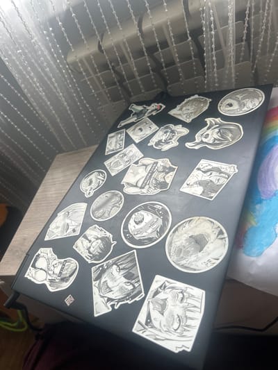The image shows a black laptop with various anime-style stickers applied to its surface. The stickers depict characters in a black and white, manga-like art style. The laptop is open and placed on a wooden surface. In the background, a white curtain with vertical beaded strands hangs over what appears to be a window or radiator. To the right of the laptop, a child's drawing of a rainbow is partially visible. The lighting suggests an indoor environment, possibly a room. There is no discernible information in the image to indicate a specific location within the city of Супонево, Russia.