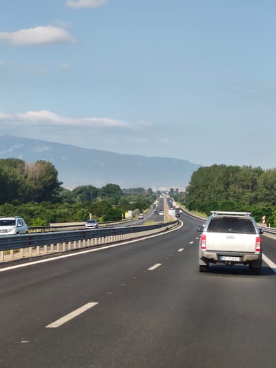The image shows a highway with traffic on a sunny day. The highway has multiple lanes and is bordered by trees on both sides. In the foreground, a silver pickup truck with the license plate "BT 1930 KT" is visible, driving in the right lane. Other vehicles are present on the highway, with some in the distance. In the background, mountains are visible under a clear blue sky with scattered clouds. The location is identified as the city of Dolni Bogrov, Bulgaria.