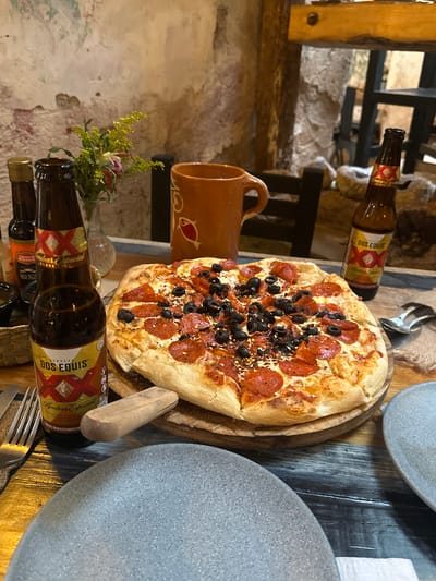 A high-angle medium shot captures a table setting with a pizza and two bottles of Dos Equis Amber Especial beer, likely in a restaurant in Zacatecas City, Mexico. The pizza, topped with pepperoni and black olives, sits on a wooden board. One bottle of beer is in the foreground to the left, and another is to the right, with its label visible. A rustic orange mug is positioned behind the pizza. The table's surface is a distressed wooden plank. The background features a rough, textured wall, a hint of greenery in a vase, and a glimpse of dark wooden furniture. The lighting is warm and inviting.